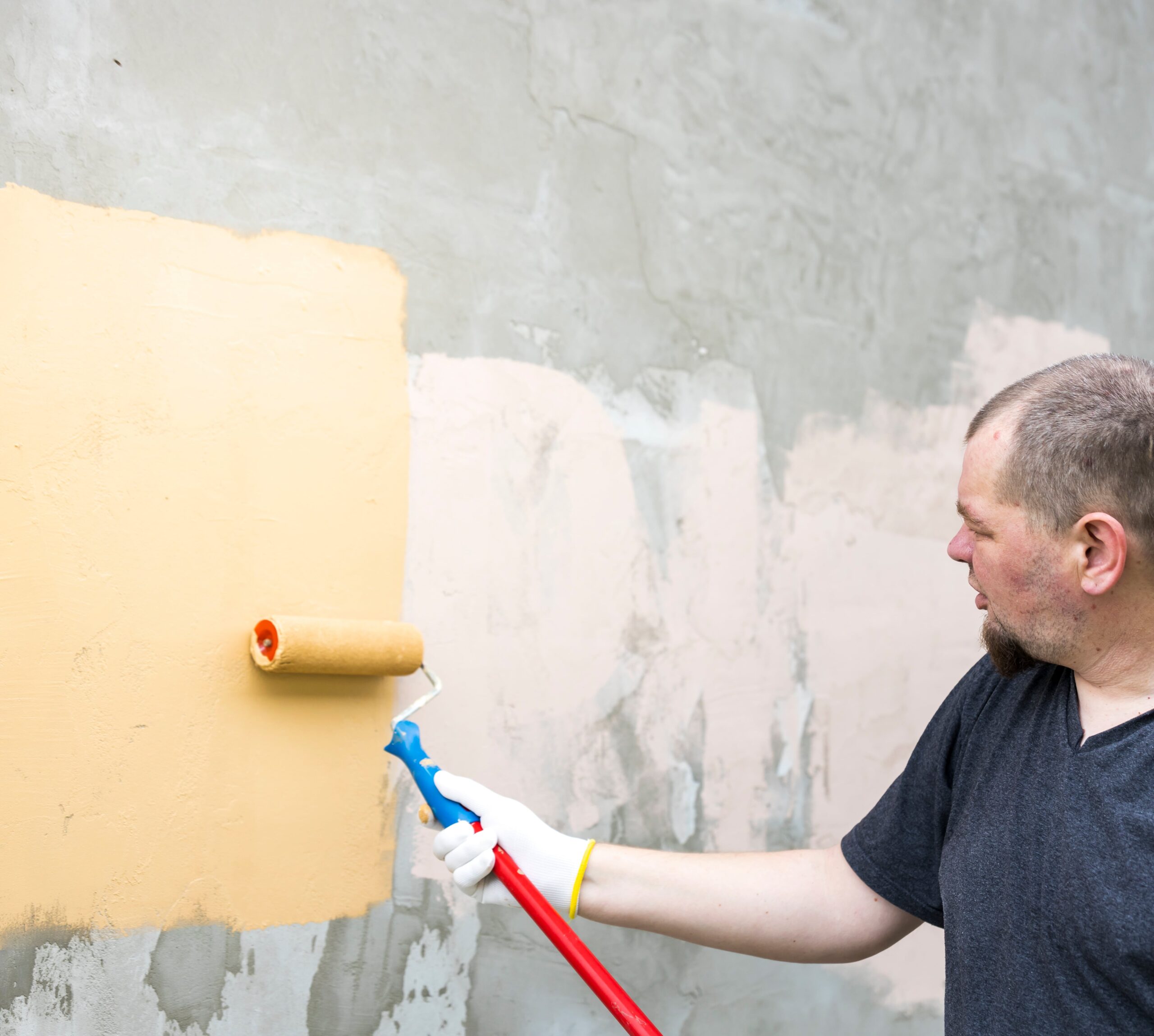 A man is painting a wall with a roller. He is wearing a black shirt and white gloves
