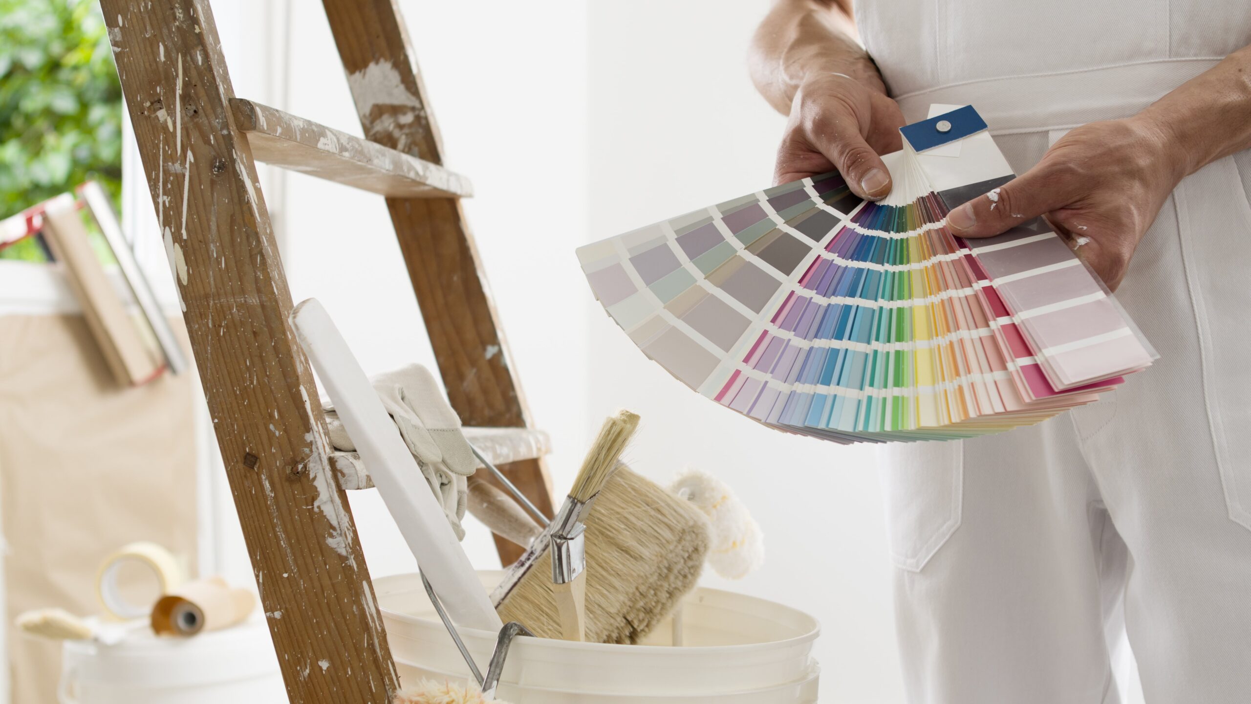 hands of house painter man decorator choose the color using the sample swatch, work of the house to renovate, a wooden ladder with paint brushes and a bucket as a background, close up